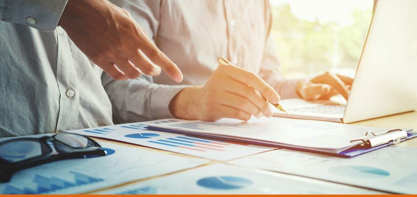 pair of employees working at a table with graphs and reports, one person is pointing at the report the other person is on the laptop.