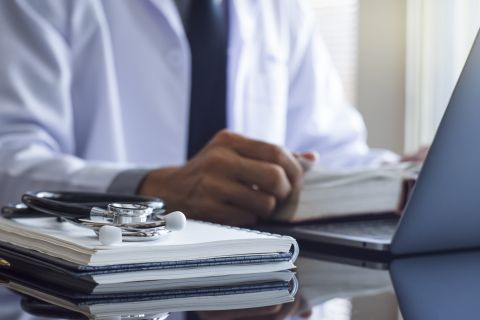 Close up of stacked notebooks with a stethoscope on top next to a hand resting on a book by a laptop